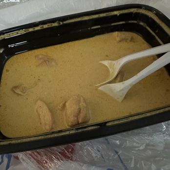 Bhetghat - Chicken Korma - A rectangular black plastic tray holds chicken korma, a dish with a yellow, creamy sauce and several pieces of chicken. Two white plastic spoons are placed inside the tray, partially submerged in the sauce. The tray is set on a surface covered with plastic and paper wrapping, indicating it may be takeout or delivery food. - Bars Near Me - Irving, Texas
