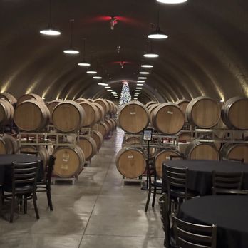 Oak Mountain Winery - Cave - A wine cellar features a tunnel with arched ceilings, lined with multiple wooden wine barrels stored on metal racks. The space is illuminated by ceiling lights, and there are tables with black tablecloths and chairs in the foreground, suggesting a tasting or event area. A Christmas tree is visible at the far end of the tunnel. - Wineries Near Me - Temecula, California