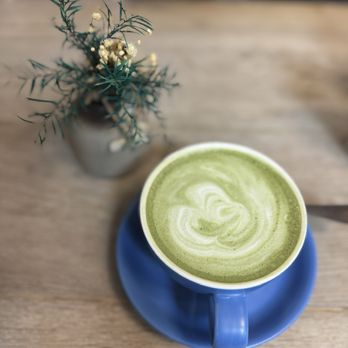 Ray's Luncheonette - Matcha latte - A photograph features a green matcha latte with a swirled pattern on its surface, served in a blue ceramic cup placed on a matching blue saucer. The cup is positioned on a wooden table. In the background, there is a small vase containing a sprig of greenery and tiny white flowers, slightly out of focus. The setting appears to be a café or casual dining area. - Breakfast & Brunch Near Me - Montclair, New Jersey