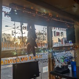 Looking out the window at sunset from my seat - A window decorated with string lights and dried chili peppers hanging in front of it. The sunset outside is visible, with trees and power lines in the background. Inside, there is a computer monitor, a black cloth, and a blue cup on a table. The window has handwritten text and stickers, indicating a business or restaurant setting.
