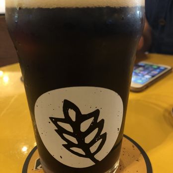 Leaven Brewing - Quaker Goats - A tall, clear glass filled with dark beer, topped with a foamy head, displays a black leaf logo on its front. The glass is placed on a yellow table with a circular coaster underneath. In the background, part of a person and a smartphone are visible, indicating a casual dining or drinking environment, likely at a brewery or pub. - Breweries Near Me - Riverview, Florida