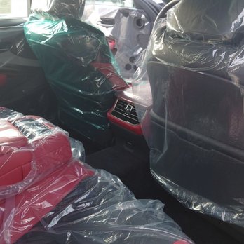 Earnhardt Lexus - The image shows the interior of a new vehicle, with plastic covers on the seats, headrests, and other surfaces. The front seats are covered with transparent plastic, and the rear seats are also wrapped in plastic. The vehicle appears to be in a dealership or showroom setting, with other vehicles visible outside the windows. The interior features red and black upholstery, and the dashboard and rear air vents are visible. - Car Dealers Near Me - Phoenix, Arizona