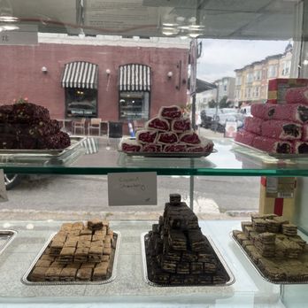 Apricot Nuts - A glass display case contains various Middle Eastern sweets such as baklava, Turkish delight, and other confections. The desserts are arranged on metal trays, with some labeled as coconut strawberry, rose petal coated pomegranate, and others. The background shows a street scene with buildings and parked cars outside the shop. - Desserts Near Me - Westfield, New Jersey
