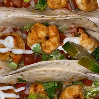 Nash & Smashed - Shrimp tacos - Three shrimp tacos are shown, each filled with grilled shrimp, diced tomatoes, onions, cilantro, and drizzled with white sauce. The tacos are served in soft flour tortillas, garnished with fresh cilantro and lime wedges. The ingredients are arranged to showcase the seafood and fresh toppings, with a focus on the vibrant colors and textures of the dish. - Sandwiches Near Me - Silver Spring, Maryland
