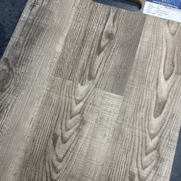A sample of luxury vinyl flooring with a wood grain pattern, featuring a textured surface that mimics natural wood. The sample has a handle cutout at the top for easy handling and a label in the top right corner providing product information, including material, size, and waterproof features. The flooring is placed on a dark surface, highlighting its light, natural wood tones.