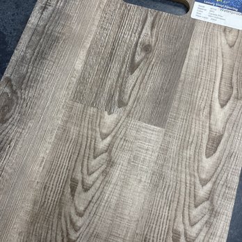 LJ Stone - A sample of luxury vinyl flooring with a wood grain pattern, featuring a textured surface that mimics natural wood. The sample has a handle cutout at the top for easy handling and a label in the top right corner providing product information, including material, size, and waterproof features. The flooring is placed on a dark surface, highlighting its light, natural wood tones. - Building Supplies Near Me - Upland, California