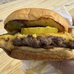 Depression Burger - A cheeseburger with a beef patty topped with melted cheese, pickles, and mustard, served in a soft bun. The burger is held in a person's hand, with a wooden surface in the background. The bun appears toasted, and the ingredients are visibly layered.