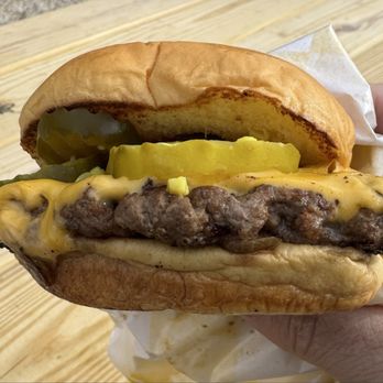 Flobee's Roadside Stand - Depression Burger - A cheeseburger with a beef patty topped with melted cheese, pickles, and mustard, served in a soft bun. The burger is held in a person's hand, with a wooden surface in the background. The bun appears toasted, and the ingredients are visibly layered. - Fast Food Near Me - Ridgefield, Connecticut