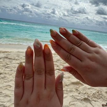 Glow Nails Salon - Perfect beach vacation nails! - Two hands with colorful, decorated nails are held up in front of a beach scene with sand, turquoise water, and cloudy sky. - Nail Salons Near Me - Chicago, Illinois