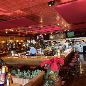 Silverado - Bar area. - The image shows the interior of a restaurant decorated for the holiday season, with poinsettia plants and festive decorations. The bar area has a curved wooden counter with red stools, and a person is working behind the bar. The ceiling has decorative panels and hanging lights, and there are Christmas ornaments hanging from the ceiling. The kitchen is visible in the background with staff preparing food, and a menu board is mounted on the wall. The overall setting is warm and inviting, with a mix of wood and brick elements. - American Near Me - Annandale, Virginia