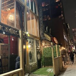 A restaurant with outdoor seating area, illuminated string lights, and signage is visible on a city street at night, with pedestrians walking by.