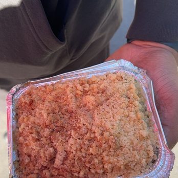 Jericho Cider Mill - Apple crisp - A person wearing a gray jacket is holding a small, rectangular foil container filled with apple crisp, a dessert with a crumbly topping. The background shows an outdoor setting with sunlight and a shadow on the ground. The focus is on the dessert and the person's hand. - Fruits & Veggies Near Me - Jericho, New York
