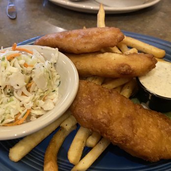 O'Niell's - A "few fries" with the battered  fish .. - A plate features two battered fish fillets, French fries, and a small bowl of tartar sauce. A side of coleslaw with shredded cabbage and carrots is also present. The dish is served on a blue plate, with a lemon wedge visible on the side. The setting appears to be a casual restaurant with a wooden table surface. - Irish Near Me - Albuquerque, New Mexico
