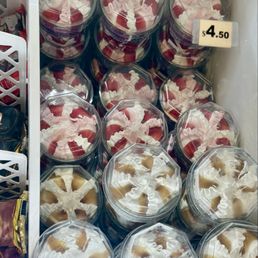 A collection of clear plastic containers filled with strawberry and caramel desserts are arranged in a freezer. The containers have transparent lids, showing the swirled whipped cream and fruit toppings inside. A small price tag indicates the cost as $4.50. The containers are stacked in an organized manner, with some on top of others, and the background includes part of the freezer interior and a white basket on the side.