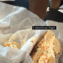 Everything bagel with veggie cream cheese. Iced Latte with oat milk. - A bagel sandwich filled with cream cheese and chopped vegetables is wrapped in parchment paper on a table. Two iced coffee drinks in clear plastic cups with straws are visible in the background. The setting appears to be a casual dining or cafe environment.