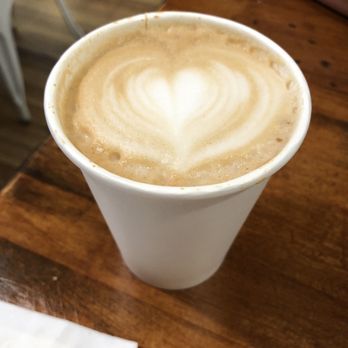 Cafe One Eight - Amaretto Latte (hot) - A hot latte with foam art in the shape of a heart is served in a white paper cup. The cup is on a wooden table, with part of a napkin visible in the foreground. The background includes a chair and a wooden floor, indicating a café environment. - Cafes Near Me - Lancaster, Pennsylvania