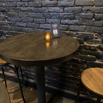 FancyFree - Seating is mostly bar stools - A round wooden table with a dark finish features a small candle holder with a lit candle and a QR code sign on top. The table is positioned against a black painted brick wall, with two wooden stools nearby. The setting appears to be in a dimly lit indoor space, likely a bar or restaurant. - Pubs Near Me - Brooklyn, New York
