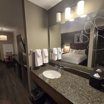 Great Wolf Lodge - The image shows a hotel room with a bathroom vanity and mirror in the foreground, displaying towels, toiletries, and a sink. The mirror reflects the bedroom with a bed, headboard, and wall art, as well as a bathroom with a toilet. The room has dark wood flooring and modern lighting fixtures. - Resorts Near Me - Mashantucket, Connecticut