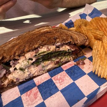 Mayfair Burger Bar - Chicken salad sandwich on rye - A toasted sandwich with visible layers of greens and creamy filling, accompanied by ridged potato chips, on a checkered paper-lined tray. - Burgers Near Me - Elberta, Michigan