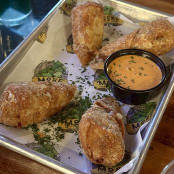 More Brewing - Italian Beef Egg Rolls - A metal tray lined with branded paper contains multiple Italian beef egg rolls, some dusted with powdered sugar or flour, accompanied by a small black cup of dipping sauce garnished with herbs. The tray is placed on a wooden table, with a glass of water visible in the background. The setting appears to be a casual dining area at More Brewing in Villa Park, Illinois, which offers craft beers and food items like egg rolls. - Brewpubs Near Me - Villa Park, Illinois