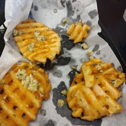 $5.49 for 4 fries - Four waffle fries topped with blue cheese crumbles are placed on parchment paper, with some crumbs and oil visible around them. The fries are golden and appear crispy, with a drizzle of sauce. The background includes a black surface and a set of keys.