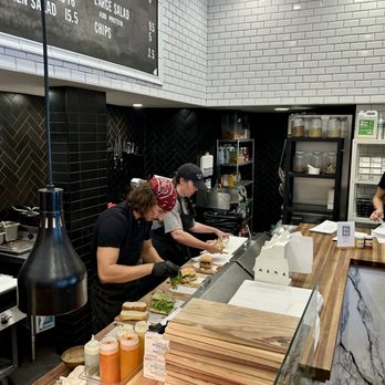 Meat & Bread - Counter - Two chefs, one wearing a red bandana and the other a black cap, prepare sandwiches behind a wooden counter in a restaurant kitchen with black and white tiled walls. - Sandwiches Near Me - Austin, Texas
