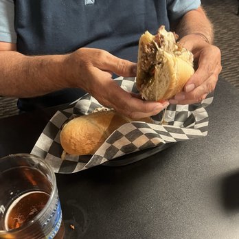 3rd Base Bar & Grill - Philly Cheesesteak Sub - A person is holding a cheeseburger with melted cheese and meat, sitting at a table with a glass of beer nearby. - Sports Bars Near Me - Olean, New York