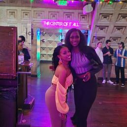 Time with friends - Two women are standing close together in a bar or entertainment venue, smiling at the camera. One woman is wearing a light-colored off-shoulder top and beige pants, while the other is dressed in a striped top and dark pants. The background includes other patrons, a neon sign that says 'THE CENTER OF IT ALL,' and colorful string lights. A television screen is visible above, displaying a talk show or interview. The setting has a lively, social atmosphere with wooden flooring and decorative wall panels.