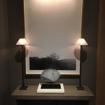 RH Denver | The Gallery at Cherry Creek - Amazing display - A decorative interior setup features a large framed black-and-white landscape photograph mounted on a wall, illuminated by a light fixture at the top. Flanking the photograph are two table lamps with shades, casting soft light and shadows. On the console table below, a stone sculpture is displayed on a stand, resting on a white base. The arrangement creates a balanced and artistic display in a modern interior setting. - Kitchen & Bath Near Me - Denver, Colorado