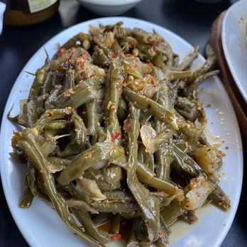 The Fly - Green beans - A white oval plate contains cooked green beans with visible seasonings, including red pepper flakes and garlic. The beans appear to be seasoned and possibly stir-fried or sautéed. In the background, there are small bowls of sauces, including a red chili sauce and a white sauce, along with other condiments. The setting suggests a meal at a restaurant or casual dining establishment. - Wine Bars Near Me - Brooklyn, New York