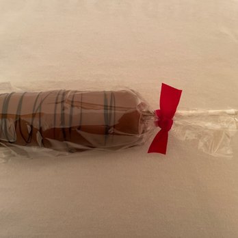 Theobroma Chocolatier - One of their beautiful milk chocolate marshmallows - A marshmallow coated in milk chocolate with a drizzle of darker chocolate on top, mounted on a stick. It is wrapped in clear plastic and secured with a red ribbon. The treat is placed on a plain, light-colored surface, likely for display or packaging. - Candy Stores Near Me - Albuquerque, New Mexico