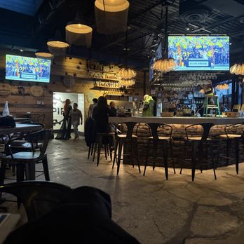 Empire - Bar area - nice floor spacing. - The photo depicts the interior of a bar or restaurant with a central curved counter, surrounded by bar stools. Hanging pendant lights illuminate the space, and two large TV screens display a scene with the label 'Ramy Brook.' Patrons are seated at tables and the bar, with some standing and walking in the background. The decor includes wooden paneling and shelves with bottles, creating a cozy yet lively atmosphere. - Burgers Near Me - Naperville, Illinois