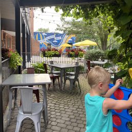 Cute outdoor seating area with giant Connect 4 for the kids - A child with blonde hair, wearing a turquoise sleeveless shirt, is playing Connect 4 outdoors on a restaurant patio. The patio features metal and plastic tables and chairs, with potted plants and flowers along the railing. In the background, there are yellow umbrellas, trees, and a mural with blue and yellow colors. String lights are hanging above the patio, and the setting appears to be a family-friendly dining area with outdoor seating.