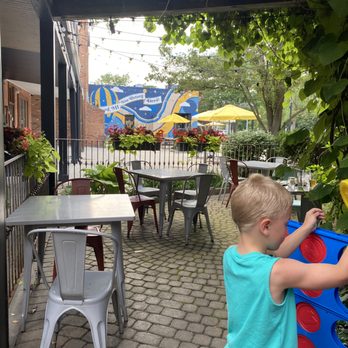Red Knapp's - Cute outdoor seating area with giant Connect 4 for the kids - A child with blonde hair, wearing a turquoise sleeveless shirt, is playing Connect 4 outdoors on a restaurant patio. The patio features metal and plastic tables and chairs, with potted plants and flowers along the railing. In the background, there are yellow umbrellas, trees, and a mural with blue and yellow colors. String lights are hanging above the patio, and the setting appears to be a family-friendly dining area with outdoor seating. - American Near Me - Oxford, Michigan