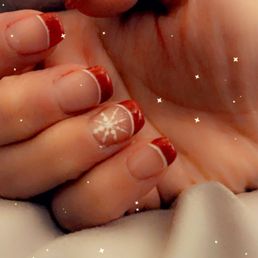 Christmas nails - The image shows a close-up of a hand with nails painted in a holiday theme. One nail displays a white snowflake design, while the others have red tips resembling a French manicure with a festive twist. The background is soft and blurred, with small star-like sparkles added for a decorative effect. The focus is on the detailed nail art, suitable for showcasing holiday nail designs.