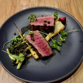 Tavernetta - Manzo ($50 - New York strip, esoterra vegetables, black garlic zabaglione [not pictured due to dairy free modification]) - The image shows a dark, round plate with two pieces of medium-rare steak, garnished with herbs and peppercorns. Accompanying the steak are grilled vegetables, including broccoli and possibly parsnip or potato, with additional garnishes and small decorative elements. The presentation is minimalist and elegant, set on a wooden table. - Italian Near Me - Denver, Colorado