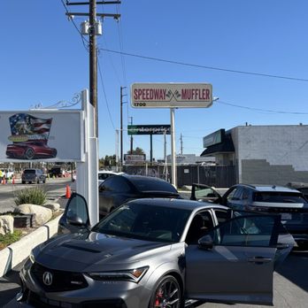 AcutintPro - A gray Acura vehicle with black wheels is parked in a lot with its front door open, near a Speedway Muffler sign and other cars. - Car Window Tinting Near Me - Upland, California