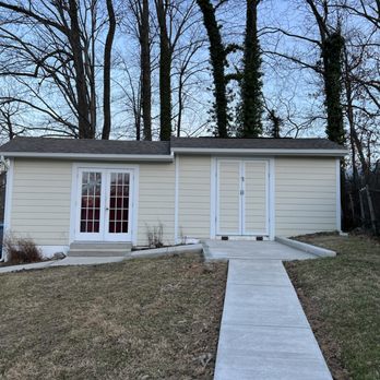 CM Hardscapes - New Concrete walkway with stone accents - A beige shed with double glass doors and a side door is positioned on a grassy yard, with a concrete walkway leading to it. Trees are visible in the background. - Masonry/Concrete Near Me