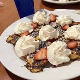 French Toast - A white plate holds French toast topped with dollops of whipped cream and sliced strawberries. The French toast appears golden brown with some darker areas, and powdered sugar is sprinkled over the dish. The plate is on a wooden table, with blue cups and other plates visible in the background, indicating a dining setting.