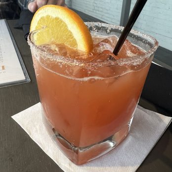 Harveys - Blood Orange Margarita - A blood orange margarita in a large, clear, square glass filled with ice and a pinkish-orange liquid. The drink is garnished with a slice of lemon on the rim. The glass is placed on a white napkin on a dark table, with a straw inserted. In the background, part of a person’s arm and a menu are visible, along with a white brick wall. - Seafood Near Me - Columbus, Mississippi