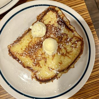 IHOP - A serving of French toast on a white plate with a blue rim, topped with two dollops of butter and sprinkled with powdered sugar. The plate is placed on a wooden table, with part of another dish visible in the top left corner. - Breakfast & Brunch Near Me - Clinton, Maryland