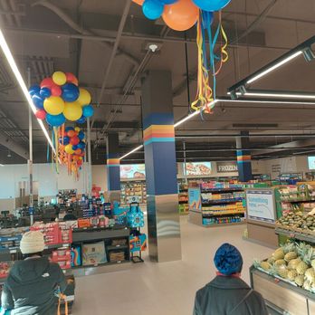 ALDI - Front of store - The photo captures the interior of an ALDI grocery store, featuring ceiling decorations including balloons and streamers in bright colors. Shelves are filled with groceries such as fruits, vegetables, and packaged goods. Two shoppers are visible, one sitting and one standing, both wearing winter hats. The store has modern lighting, and digital screens display promotional content. The overall scene suggests a festive or special event within the store. - Grocery Near Me - Washington, DC, District of Columbia