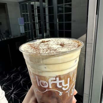 Lofty Coffee - A person holds a clear plastic cup filled with iced coffee topped with cream and cocoa powder. The cup has the logo 'lofty' printed on it. The background features a modern building with large glass windows and a gray exterior, suggesting an urban or commercial setting. - Coffee & Tea Near Me - Carlsbad, California