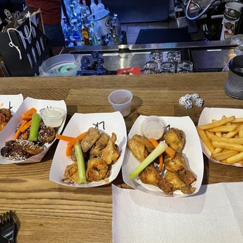 Knucklehead Pub - Wings - Four paper trays with chicken wings, fries, and vegetables are arranged on a bar counter, with a bar area and bottles in the background. - Bars Near Me - Eagle, Wisconsin