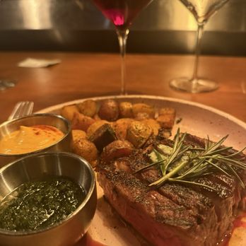 Ossobuco Miami- Steak house - A dinner plate features a grilled steak garnished with rosemary, roasted potatoes, and two small bowls of sauce, with two glasses of red and white wine behind. - Steakhouses Near Me - Miami, Florida