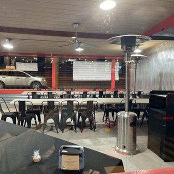 Pizza 9 - Patio sitting area - The image shows an indoor patio dining area at a restaurant, with several tables and black chairs arranged in rows. Ceiling fans and patio heaters are mounted on the ceiling. The space has a metal wall on one side and large windows with a view of parked cars and the street outside. Small decorative items are placed on the tables, and a black shelf with a 'Thank You' sign is visible in the corner. - Pizza Near Me - Grants, New Mexico