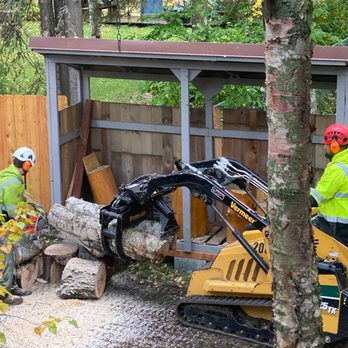 Carlos Tree Service - machine holding the logs as its being cut into firewood lengths directly in front of the empty woodshed - Tree Services Near Me - Anchorage, Alaska