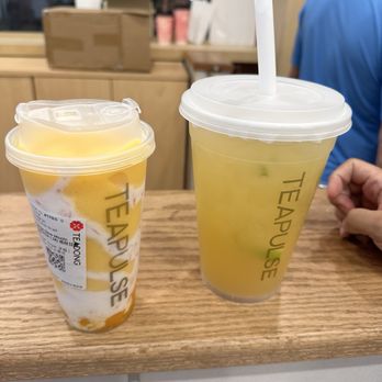 Tea Pulse - Jasmine Lemon Jumbo and Mango Dew Mochi Smoothie ( M) - Two cups of mango lemon beverages from Tea Pulse are placed on a wooden counter, one with a straw and the other with a sealed lid, inside a cafe. - Bubble Tea Near Me - Flushing, New York
