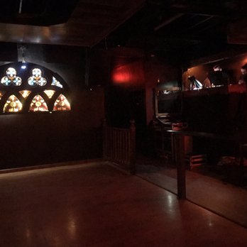 The High Dive - DJ - Dark interior of a Gothic-themed bar featuring stained glass windows and a DJ booth with two DJs or staff members. - Cocktail Bars Near Me - Hamtramck, Michigan