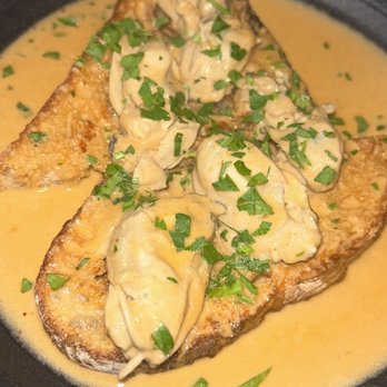 Osa - Oysters - The image shows a dish with several oysters placed on toasted bread slices, covered in a creamy, light-colored sauce. The dish is garnished with chopped green herbs, likely parsley. It is presented in a black bowl, suggesting a restaurant setting, emphasizing seafood and rich flavors. - Cocktail Bars Near Me - Middletown, Connecticut